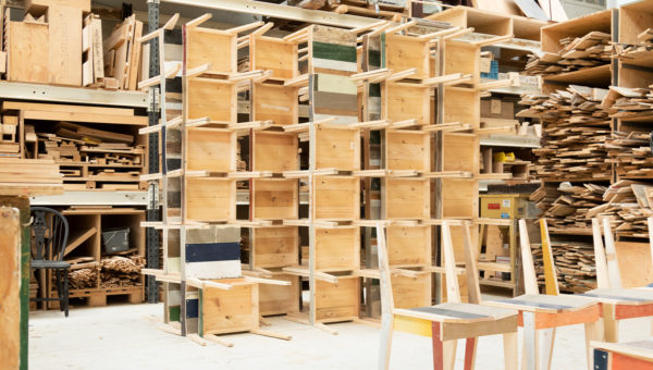 A workshop filled with wooden shelving units and various wooden materials organized on shelves, with chairs partially visible in the foreground.