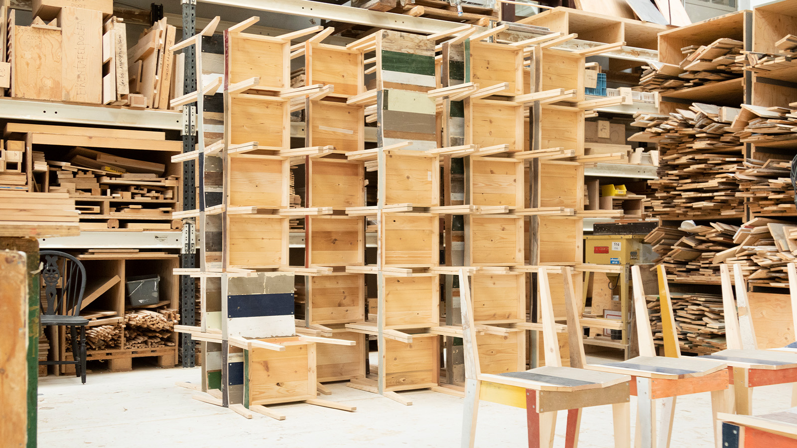 A workshop filled with wooden shelving units and various wooden materials organized on shelves, with chairs partially visible in the foreground.