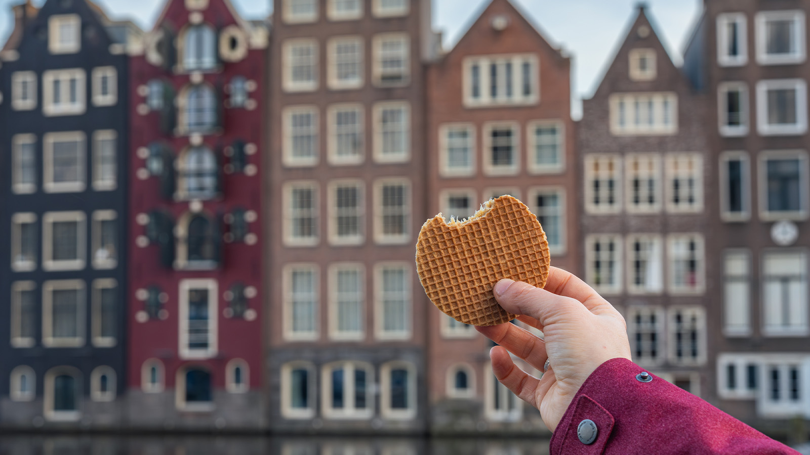 A hand holding a bitten stroopwafel in front of traditional narrow and tall Dutch buildings, with their distinctive gabled facades, in the background.