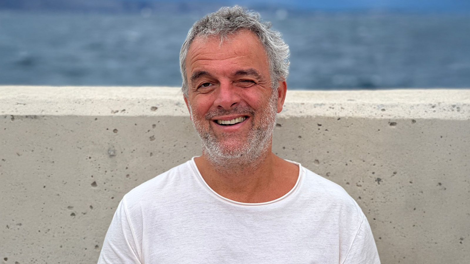 A man with gray hair and a beard wearing a white shirt stands smiling against a concrete wall, with a blurred ocean visible in the background.