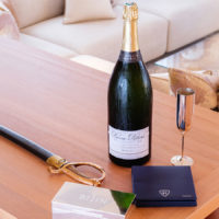 Table with a large bottle of champagne, a saber, a metallic flute, and branded items, in a luxurious living room with cream-colored sofas and decorative pillows.