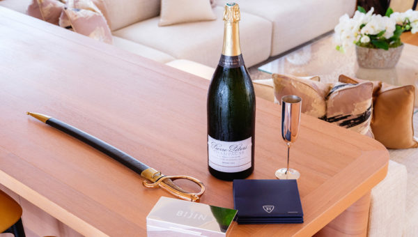 Table with a large bottle of champagne, a saber, a metallic flute, and branded items, in a luxurious living room with cream-colored sofas and decorative pillows.