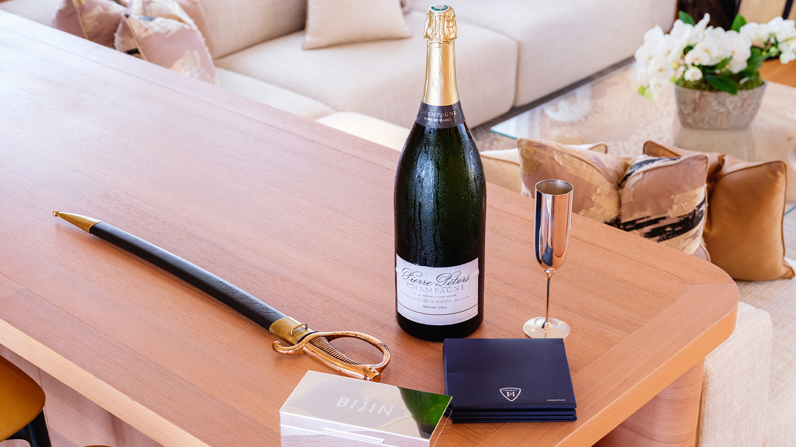 Table with a large bottle of champagne, a saber, a metallic flute, and branded items, in a luxurious living room with cream-colored sofas and decorative pillows.