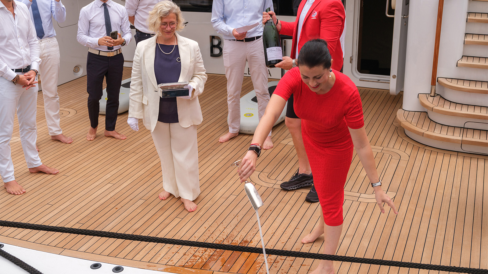 A group of people on a yacht's wooden deck participating in a champagne christening ceremony, with a woman in a red dress pouring champagne over the side while others watch.