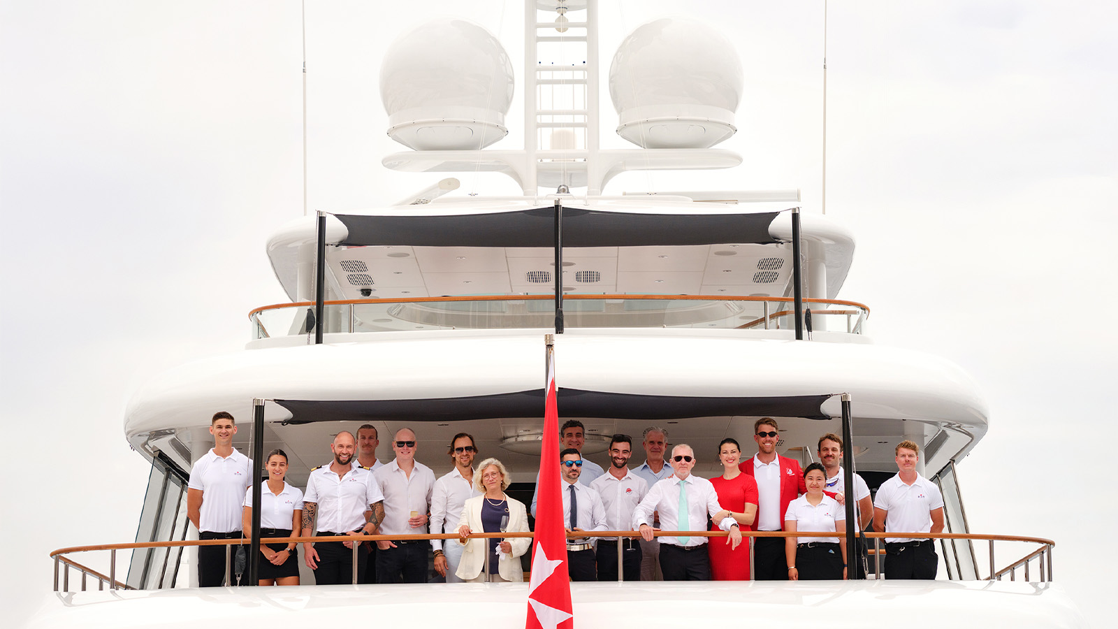 A group of people standing on the deck of a large yacht, posing for a photo. The yacht has a sleek, white design and large dome structures on its top. A red flag with a white star is in the foreground, and the group consists of men and women, some in formal attire and some in uniform.