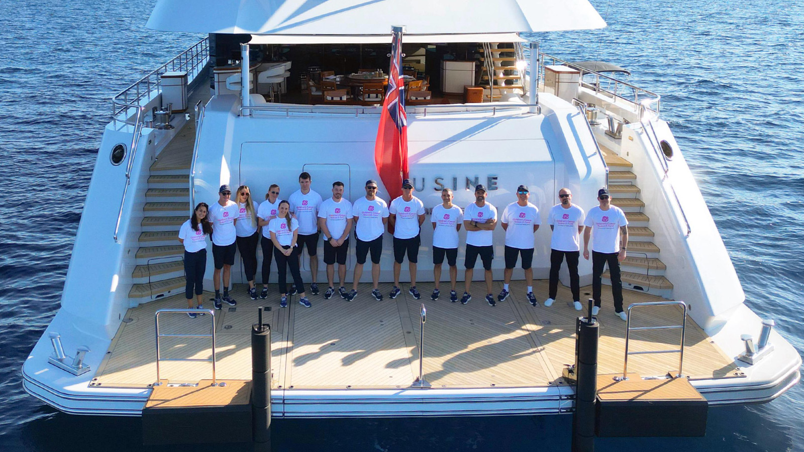 A group of people in matching white shirts and dark shorts stand on the deck of a large yacht named 'CUSINE', which is docked on the water. A red flag is hoisted at the back of the yacht, and the ocean is visible in the background.