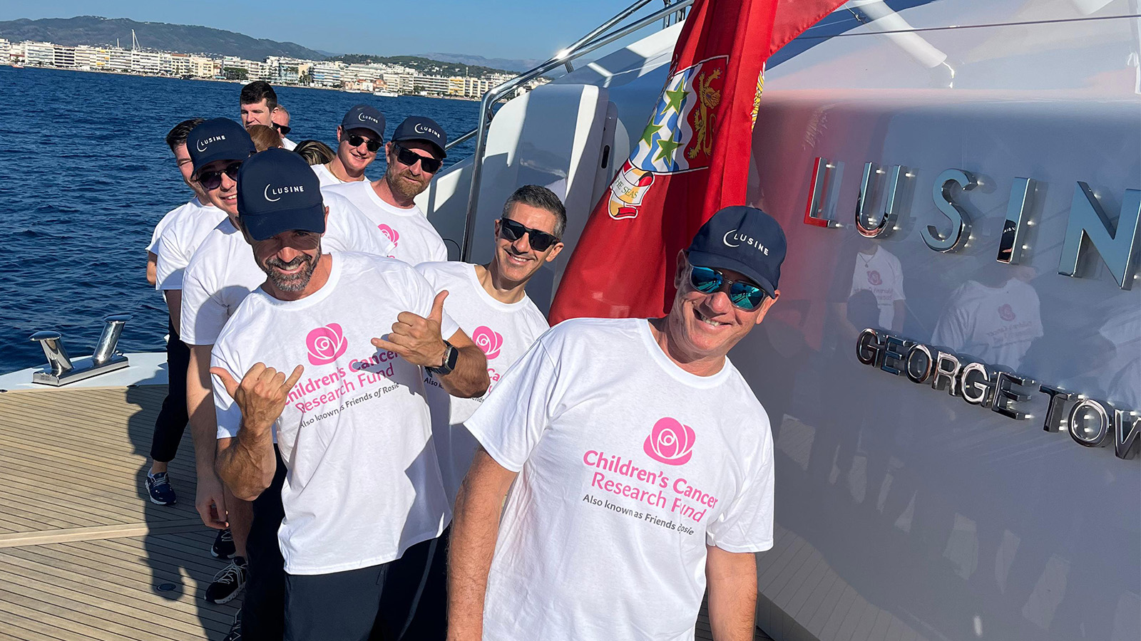 A group of smiling people wearing matching white t-shirts with "Children's Cancer Research Fund" logo and text, standing on a boat with a city and water in the background. They are wearing navy blue caps with "Lusine" written on them and posing enthusiastically for the photo.