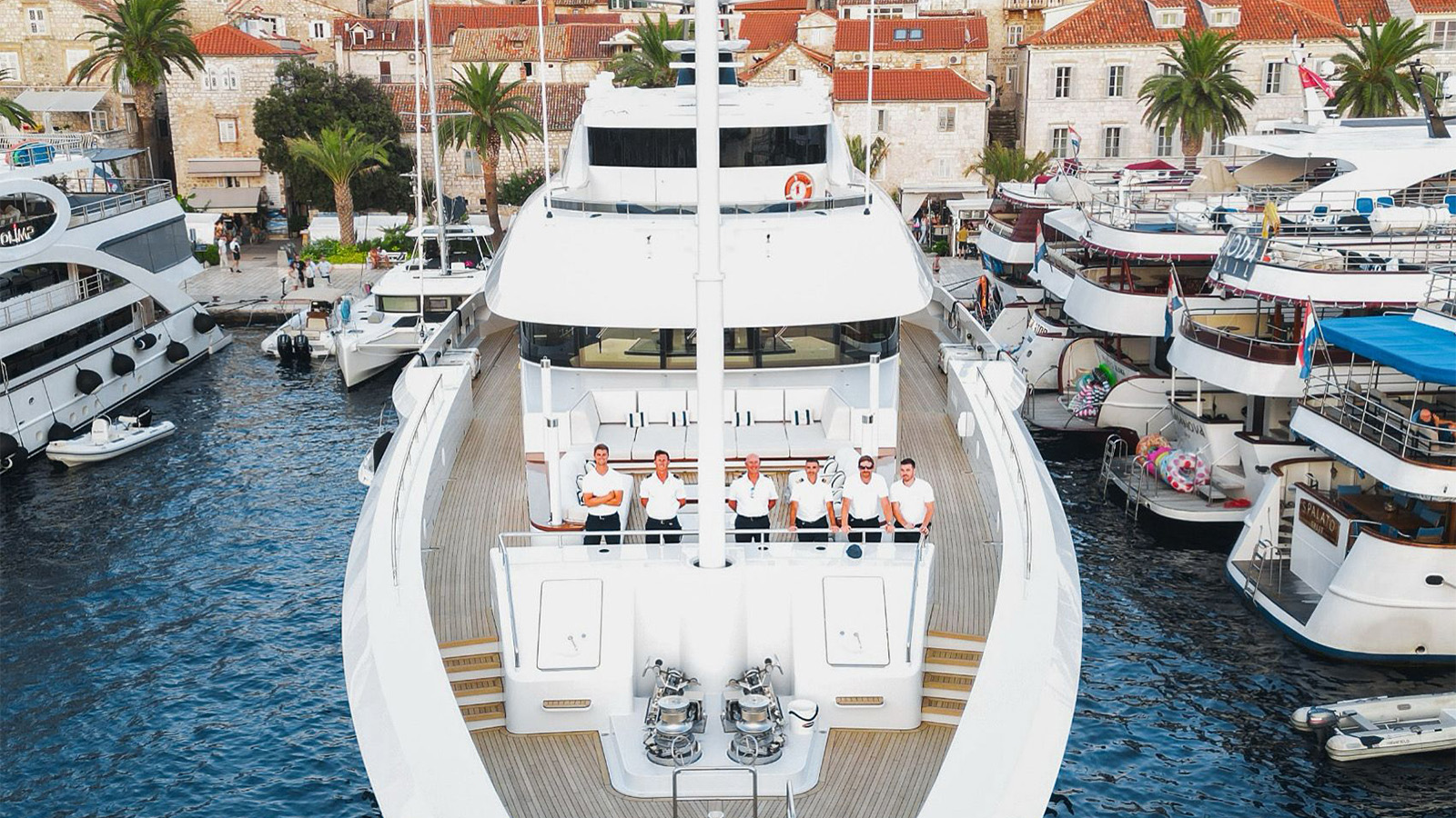 A large yacht docked at a marina with seven people in uniform standing on the deck, surrounded by other smaller boats and a backdrop of Mediterranean-style buildings with red-tiled roofs and palm trees.