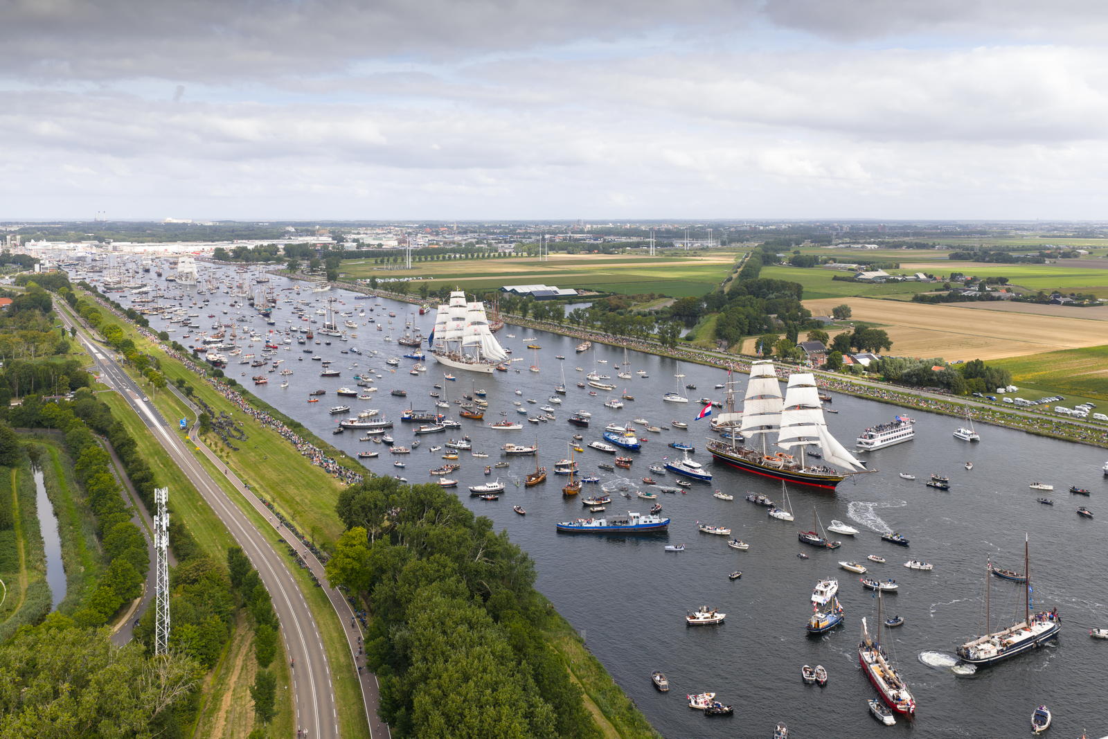 An aerial view of a busy waterway filled with numerous sailing ships, yachts, and smaller boats, next to a road lined with trees and open fields, under a cloudy sky.