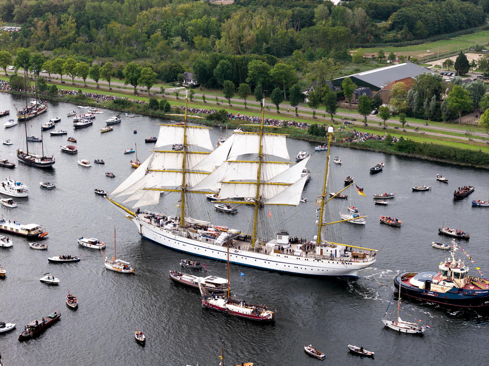 Aerial view of a large sailing ship with three masts and numerous smaller boats surrounding it on a waterway, with a green landscape in the background and crowds of people watching from the shoreline.