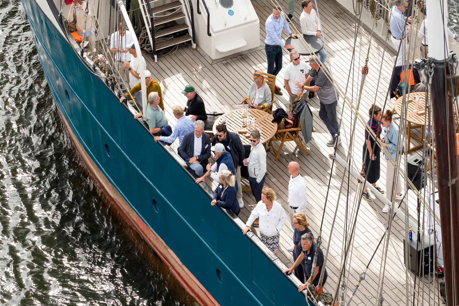 A group of people gathered on the deck of a ship, some sitting around a table and others standing near the railing, with the water visible below.