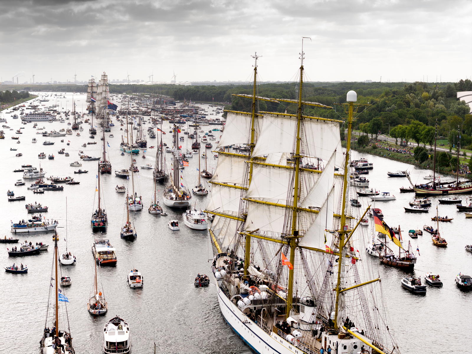 A large regatta featuring numerous sailing ships and boats crowded together on a wide river, with a prominent tall ship in the foreground. The sky is overcast, and spectators line the riverbanks to watch the event.