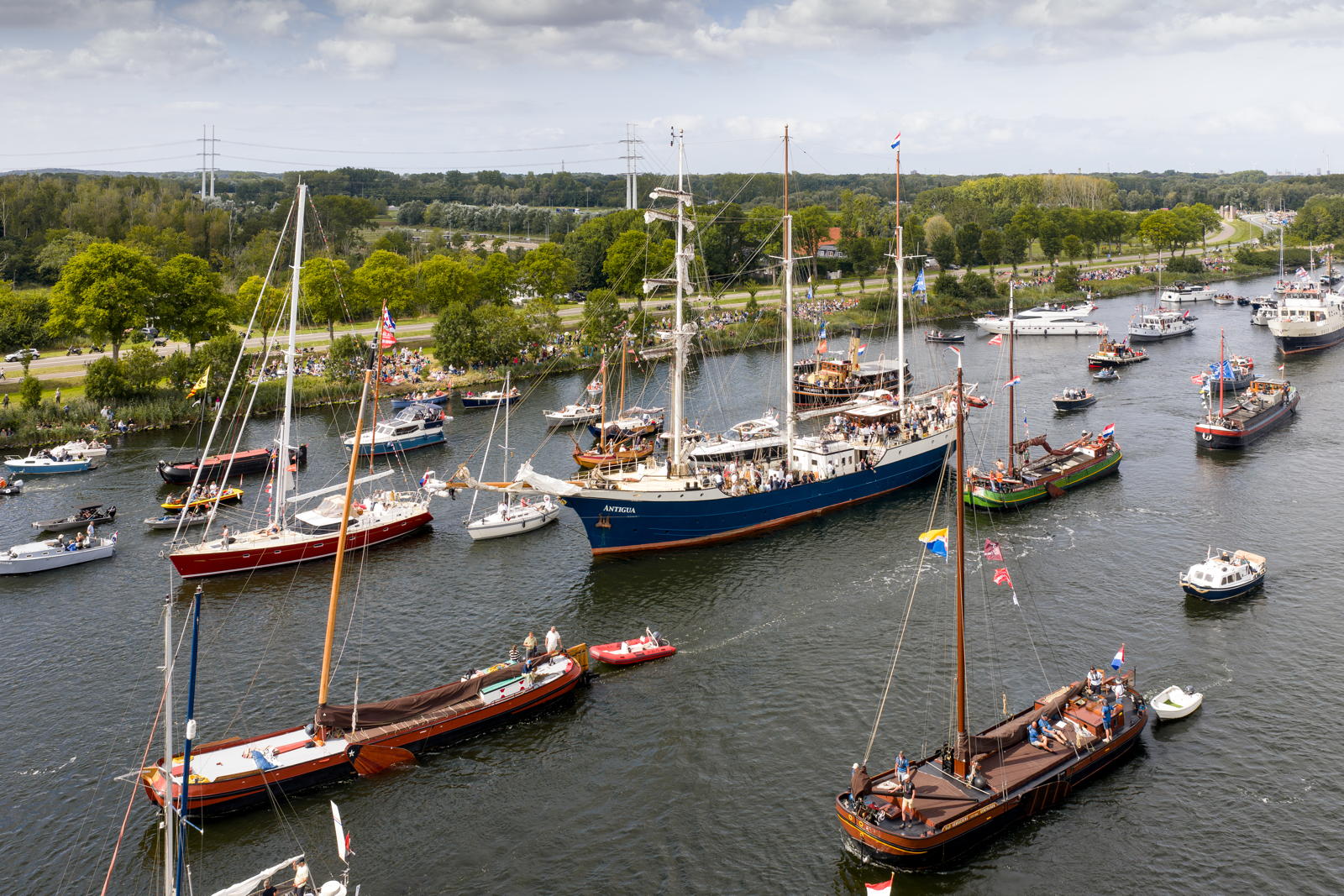 An aerial view of a busy waterway with numerous sailboats and ships participating in a maritime event. The shore is lined with spectators, and lush green trees and a road are visible in the background under a partly cloudy sky.