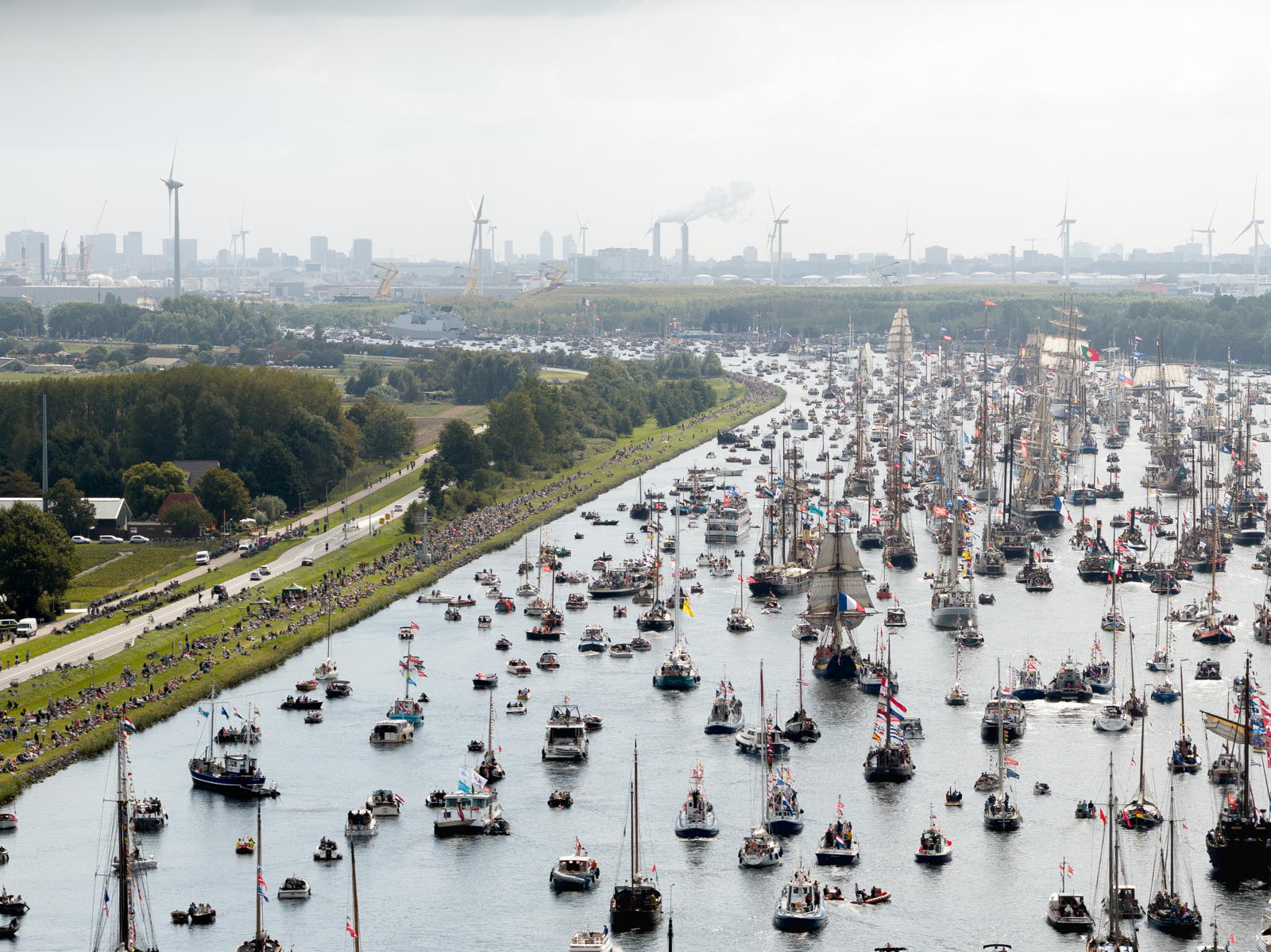 A large number of boats, including sailboats and motorboats, fill a wide canal lined with spectators. The background shows a cityscape with wind turbines and industrial structures under a cloudy sky.