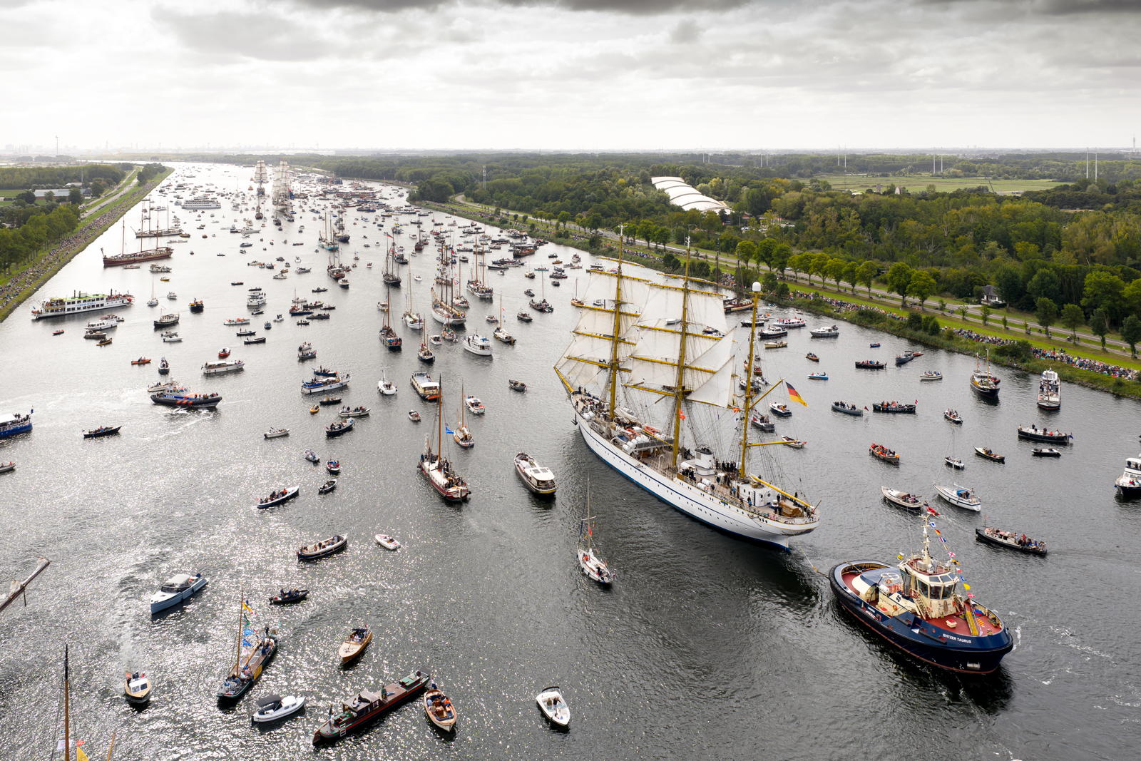 Aerial view of a large sailboat with multiple masts navigating a busy waterway filled with numerous smaller boats, surrounded by trees and spectators on the shore.