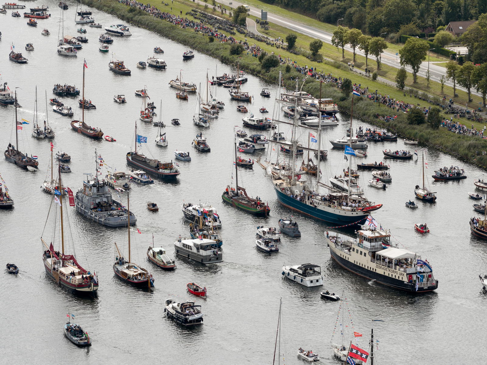 Aerial view of a busy waterway filled with a variety of boats, including sailboats and motorboats, during an event. The banks are lined with spectators, and a nearby road runs alongside the water.