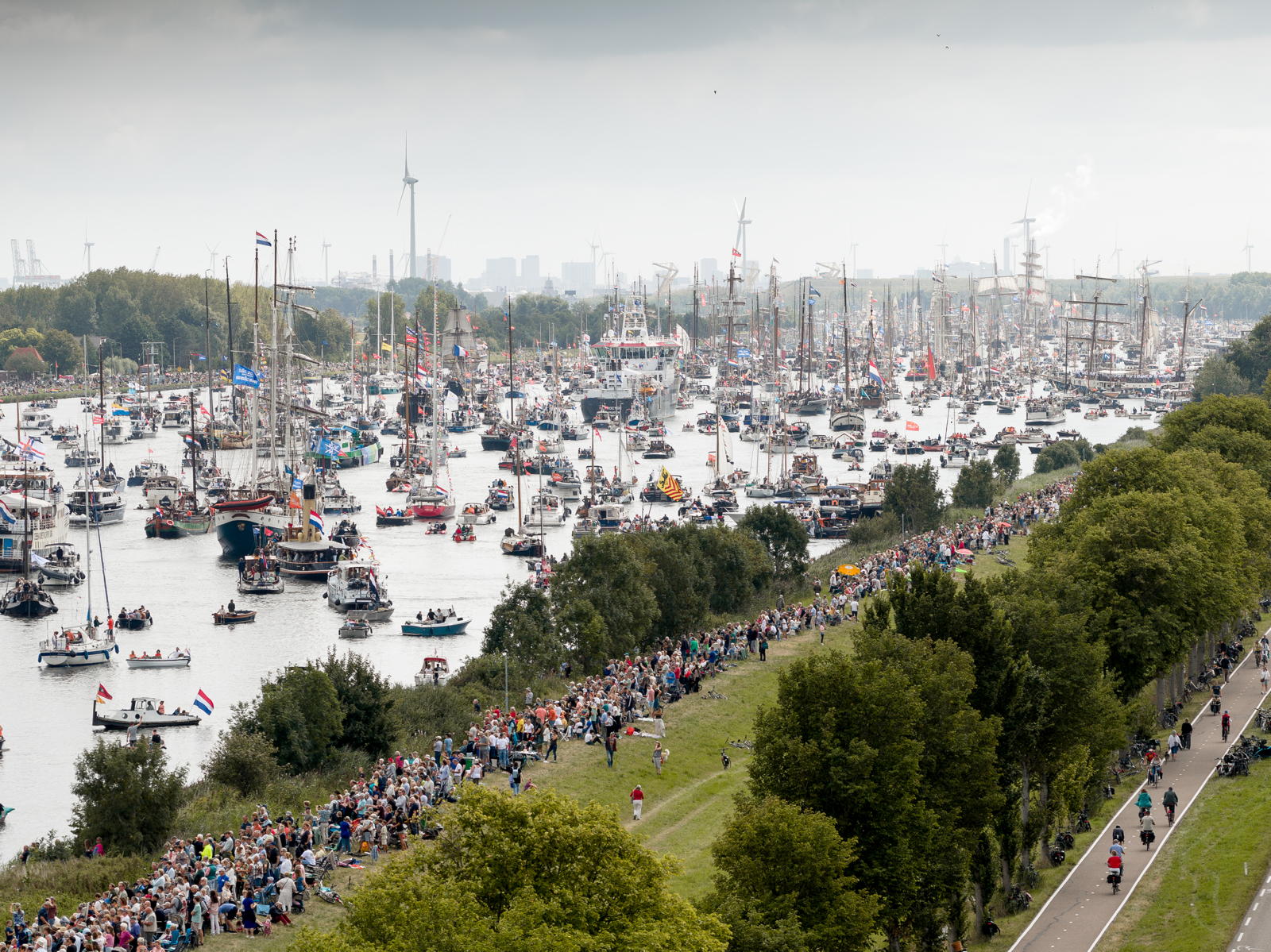 Aerial view of a crowded waterway filled with numerous sailboats and ships, with flags flying. A large number of people are gathered along the shore watching the event. There are wind turbines and city buildings visible in the background.