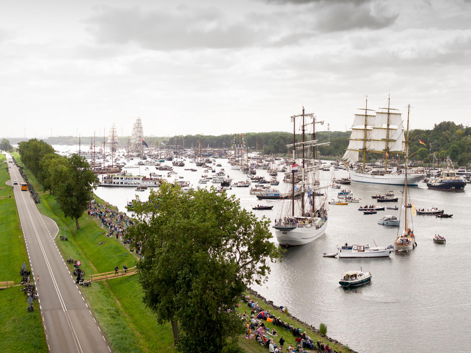 A large fleet of sailing ships and smaller boats is gathered on a wide river, with tall ships featuring masts and sails prominently visible. A road with scattered people watching the event runs parallel to the river, and the scene is set against a background of green trees and a cloudy sky.