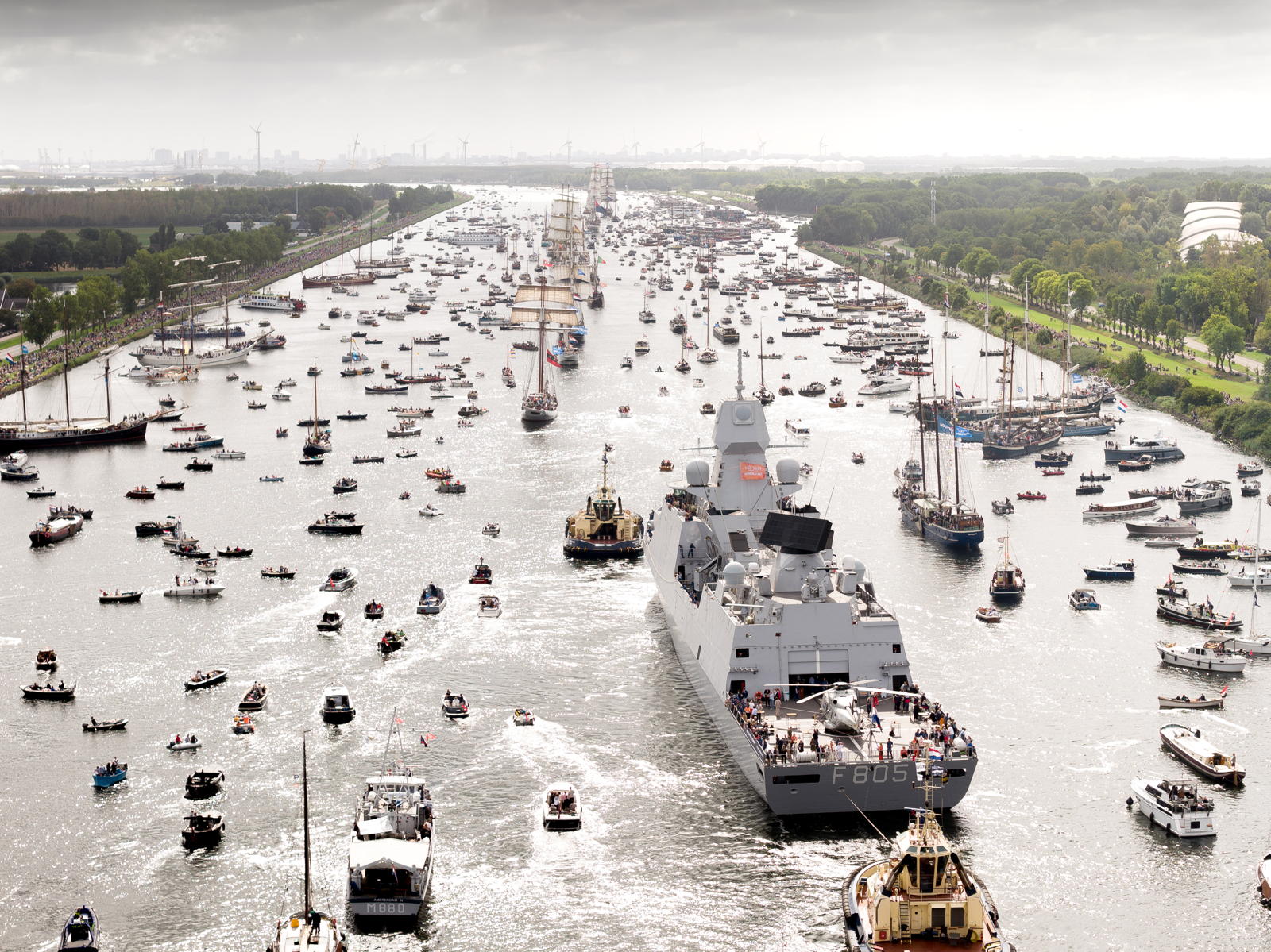 A large naval vessel marked with "F805" sails down a crowded waterway, surrounded by numerous smaller boats. The scene is lively with spectators visible on the boats and along the banks. The sky is overcast and the surrounding landscape is lush with greenery.