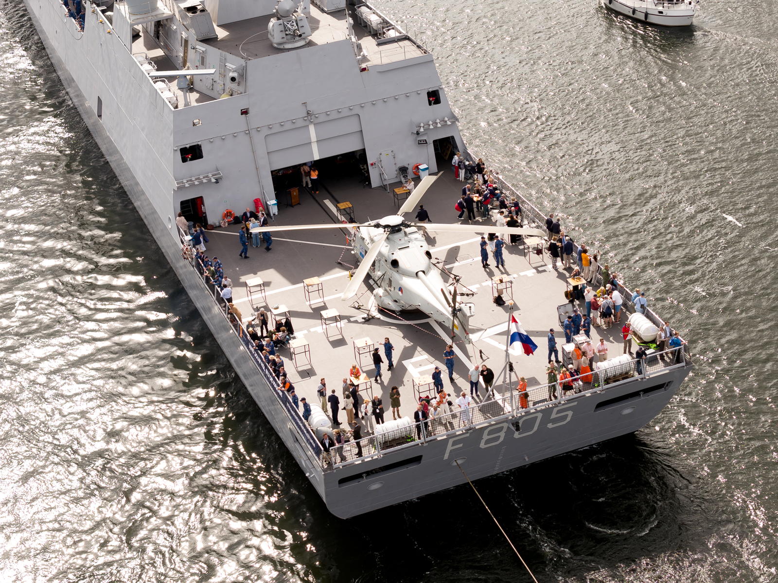 A naval ship with identification number F 805 on the deck, featuring numerous people and a helicopter. The ship is in a body of water, with a smaller boat visible in the background. A flag is prominently displayed on the rear of the ship, and the sun casts reflections on the water.