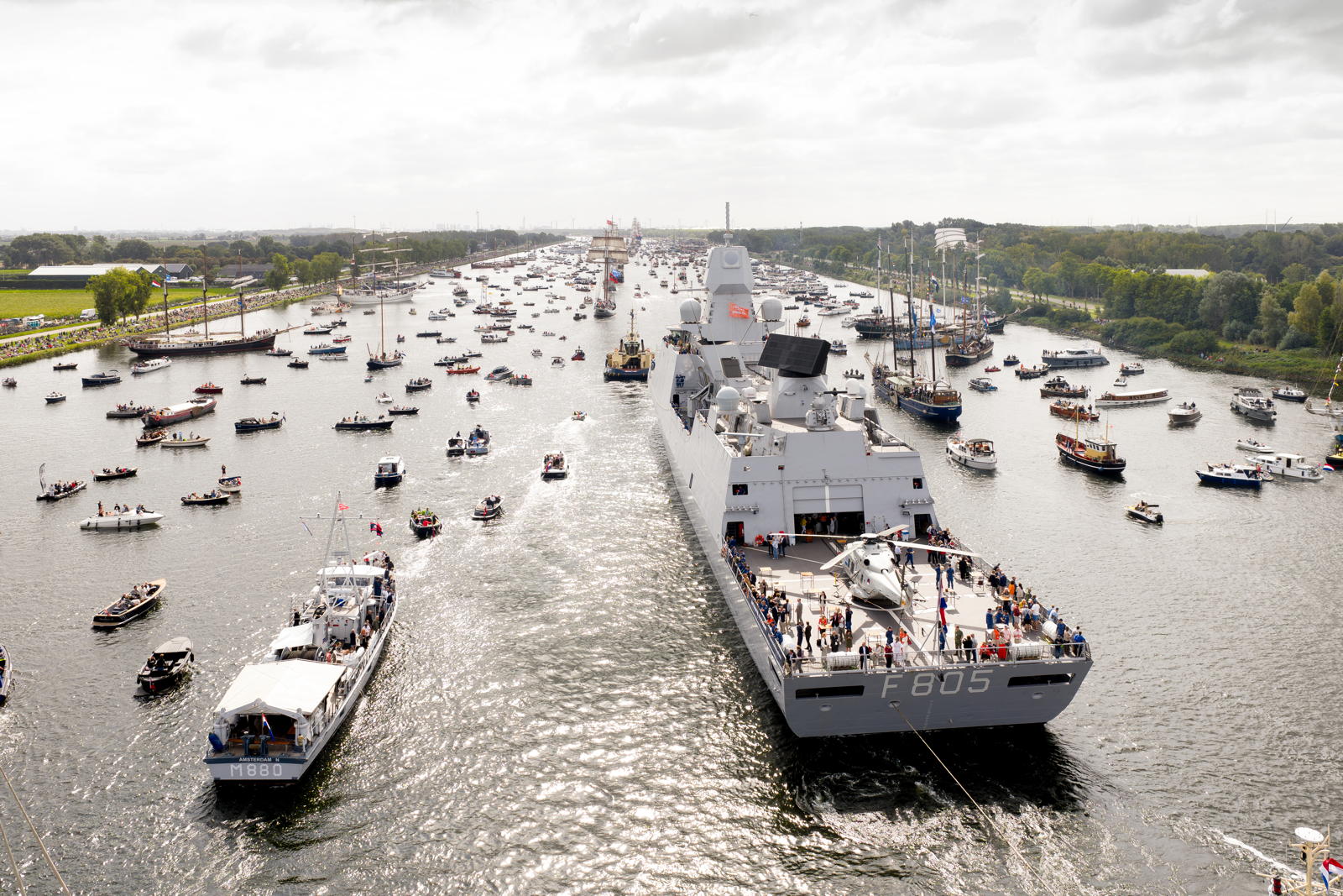 A large naval ship, identified by markings F805, navigates a crowded waterway during a maritime event, surrounded by numerous smaller boats. People are visible on the decks of the ship and some smaller boats, indicating a festive atmosphere. The waterway is lined with green vegetation and distant spectators.