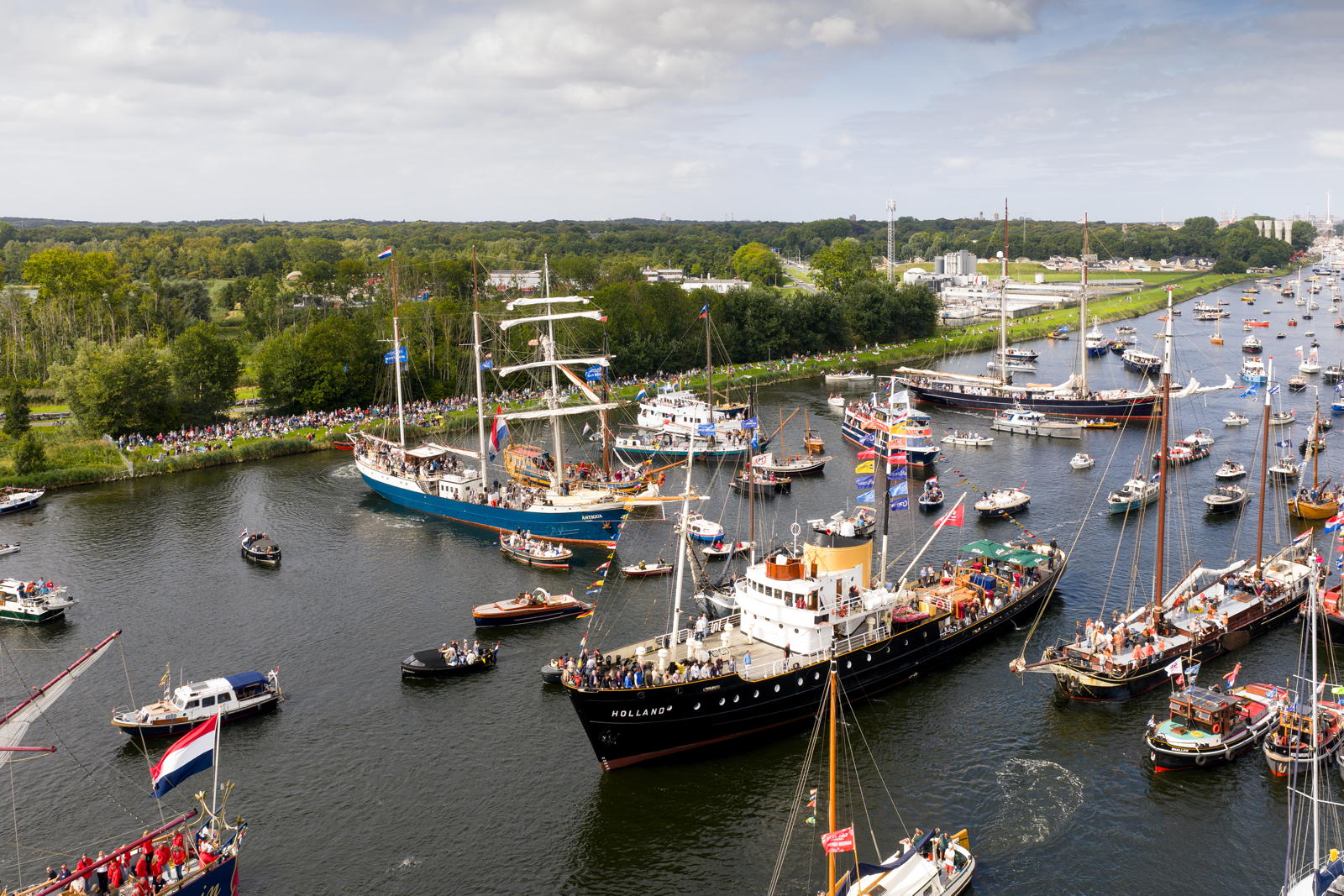 An aerial view of a busy waterway filled with numerous boats and ships decorated with flags, surrounded by lush greenery and a crowd of spectators lining the riverbank under a cloudy sky.