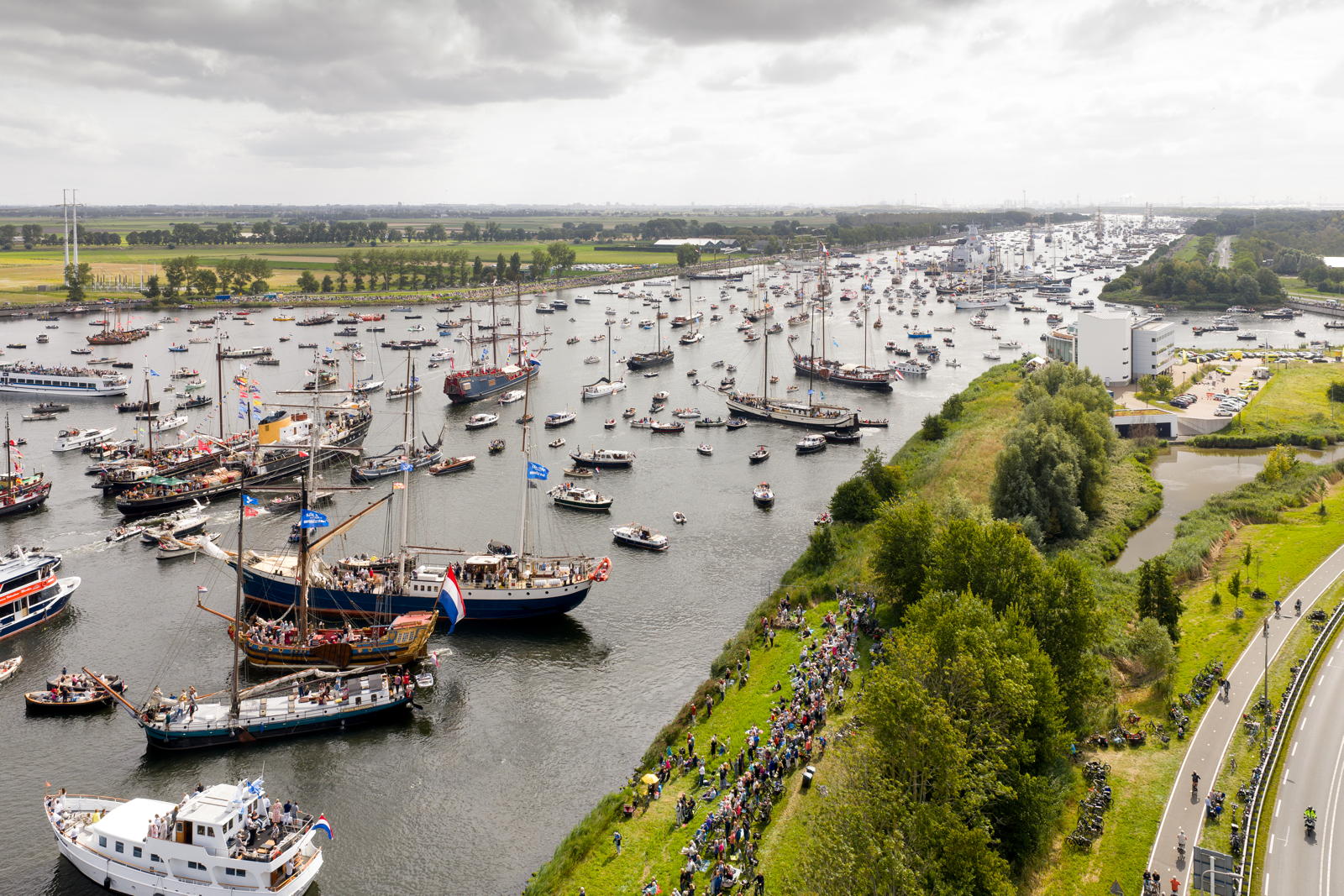 A large fleet of various ships and boats on a river, with people lining the grassy riverbank to watch the event. The sky is overcast, and the landscape is lush with greenery and distant fields.