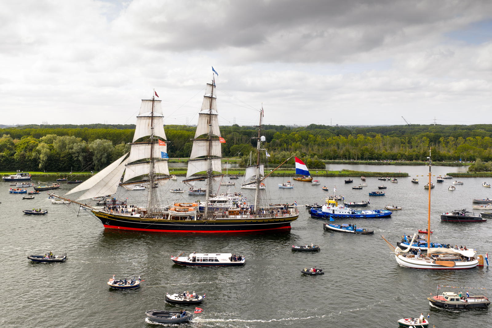 Numerous boats sail on a river with a large, traditional sailing ship featuring multiple masts in the foreground, surrounded by smaller vessels. A Dutch flag is visible on the sailing ship. The background displays lush green trees and a partly cloudy sky.