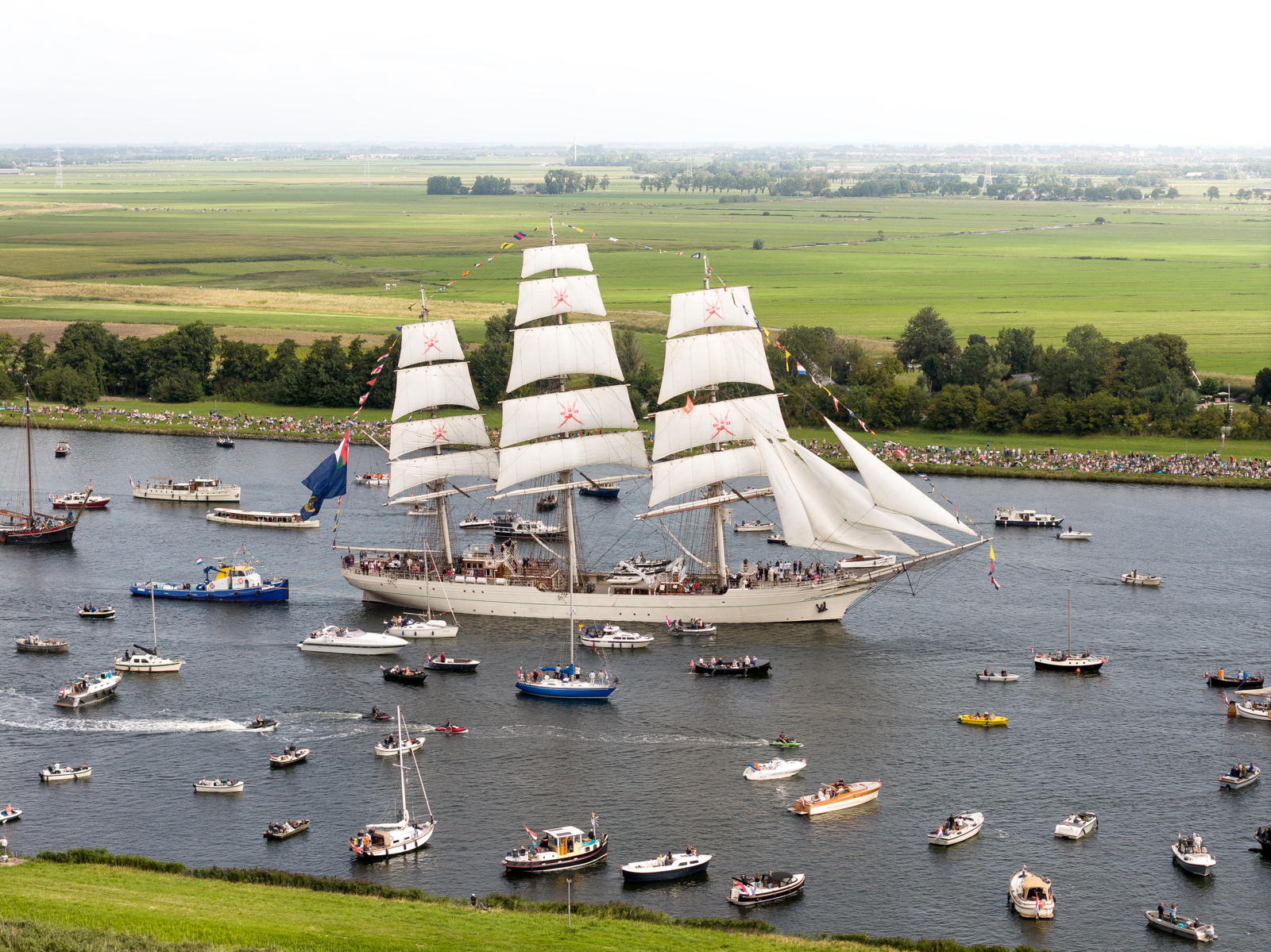 A large sailing ship with multiple white sails and colorful flags, surrounded by numerous smaller boats on a river, with spectators gathered along the grassy riverbanks in the background.