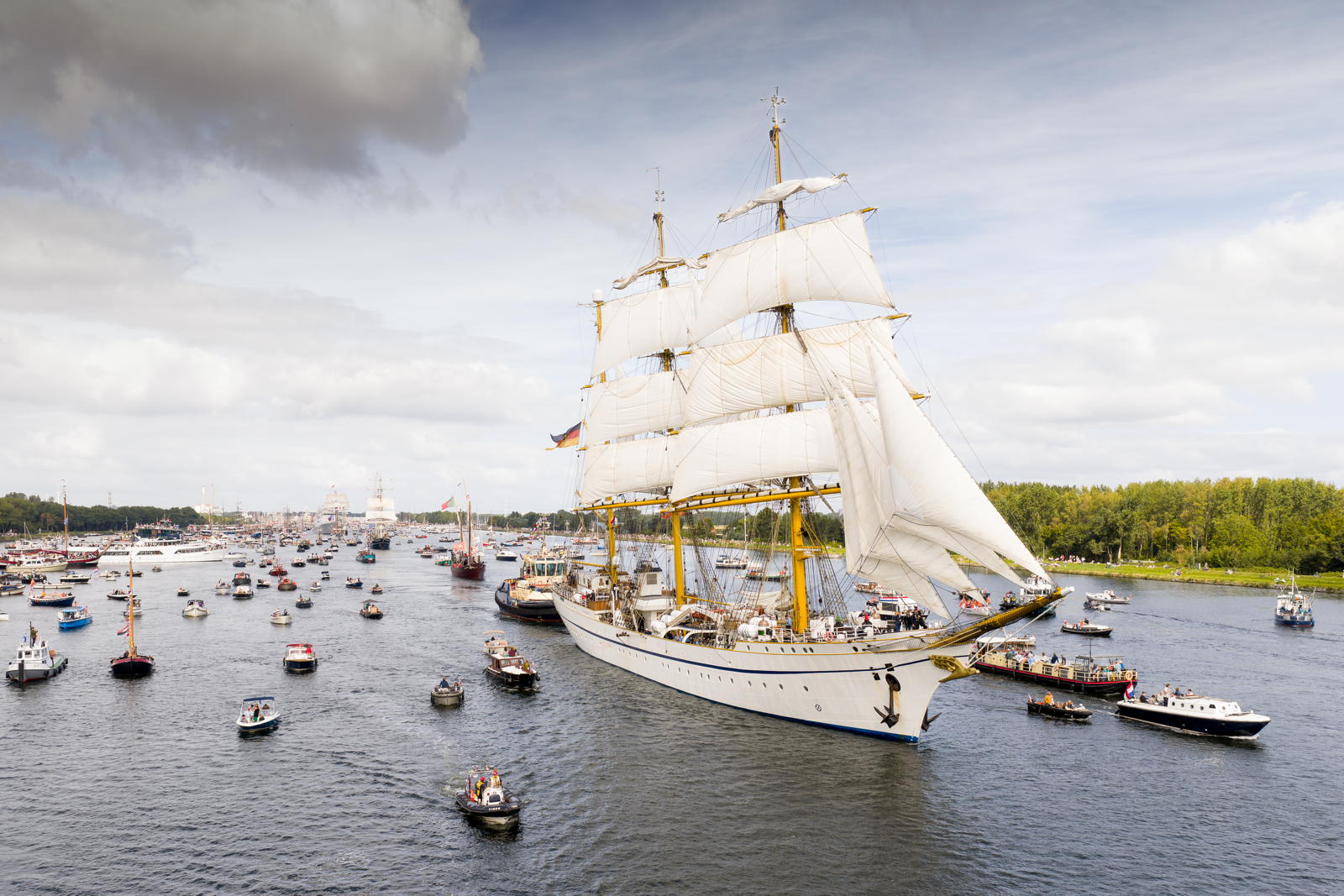 A large sailing ship with multiple white sails navigates a busy waterway surrounded by smaller boats, under a partly cloudy sky.