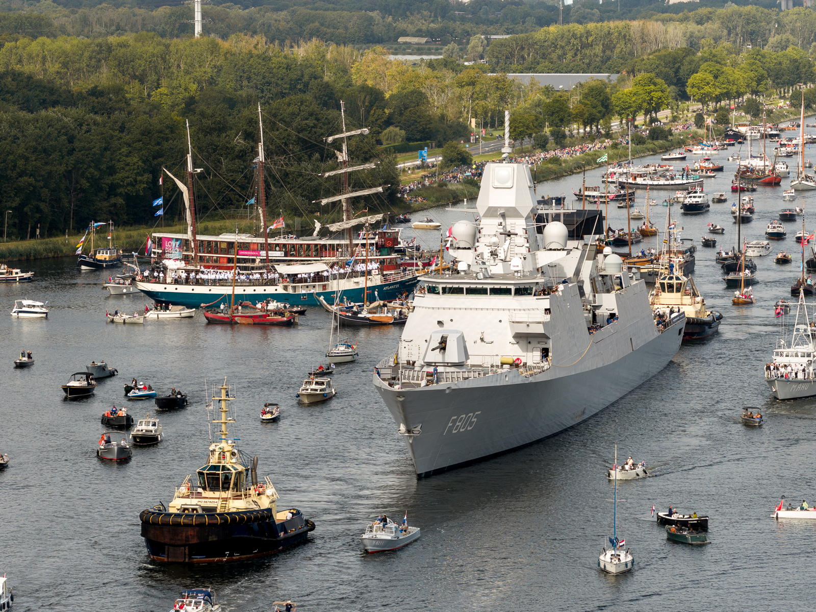 A large naval ship navigates through a crowded waterway filled with various smaller vessels, including sailboats and pleasure craft, against a backdrop of trees and spectators lining the riverbank.