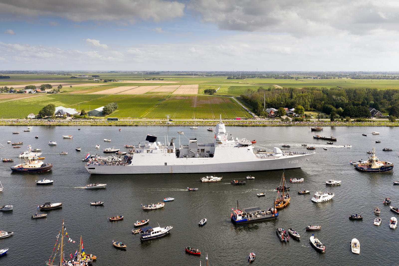 A large naval ship, marked F805, navigates through a waterway surrounded by numerous smaller boats and vessels. The landscape in the background includes green fields and a few buildings under a partly cloudy sky.