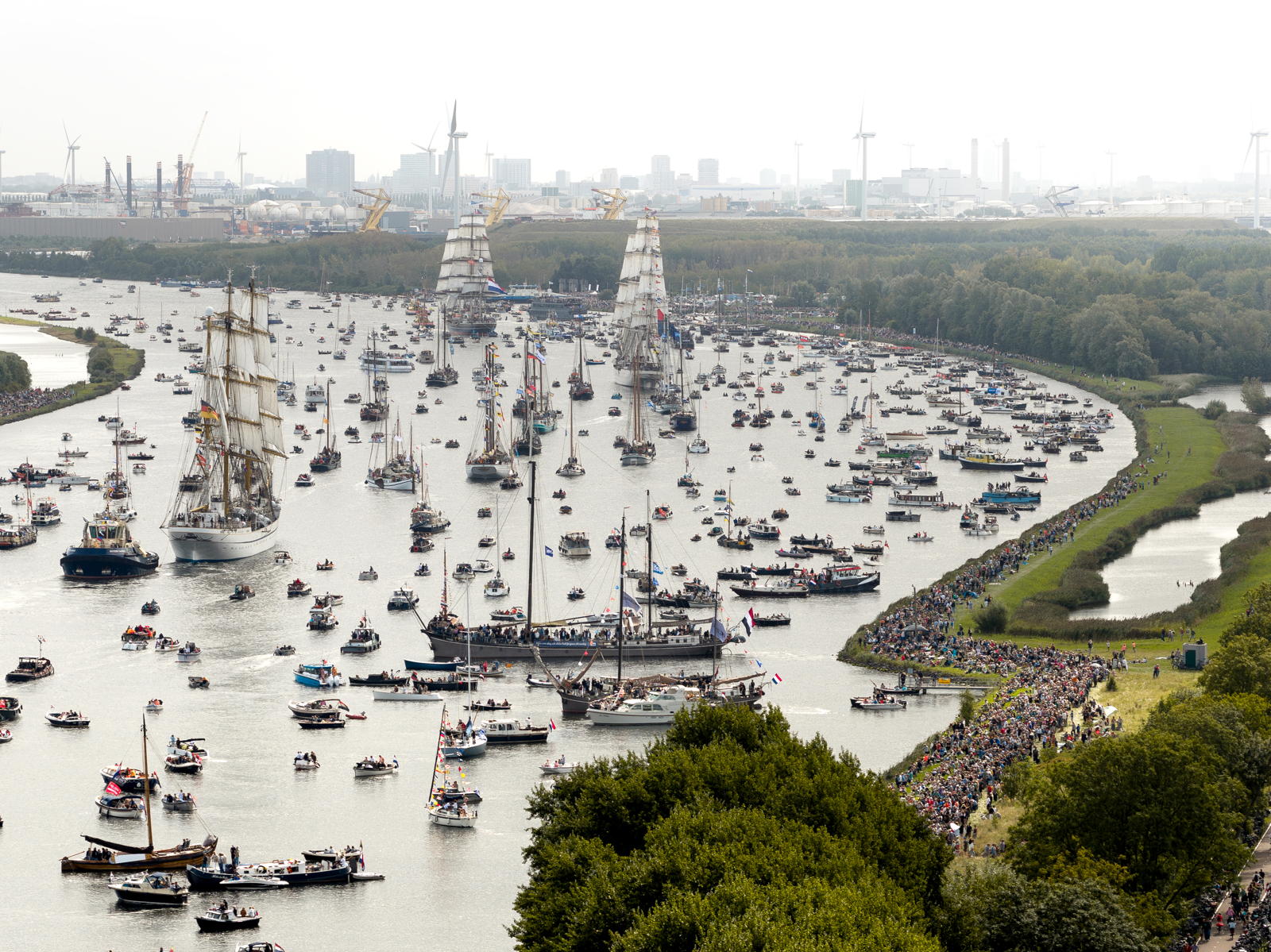 Aerial view of a crowded waterway decorated with numerous boats, including several tall ships with sails. Large crowds line the banks of the river, viewing the maritime spectacle. The background features industrial buildings and wind turbines.