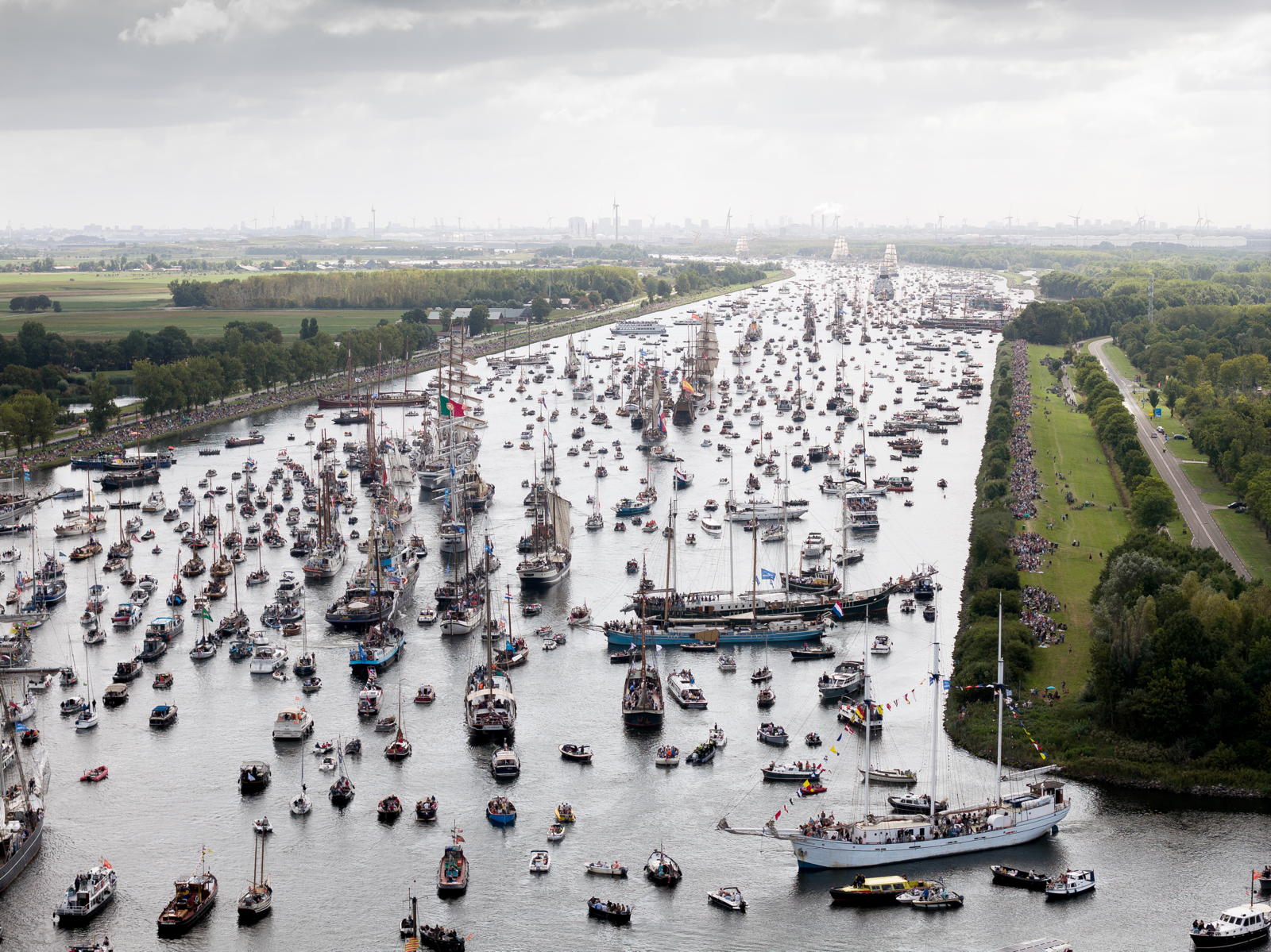 Aerial view of a crowded waterway filled with a large number of boats and ships of various sizes, with people gathered on the banks to watch. The sky is overcast, and the scene includes lush green landscapes surrounding the water.