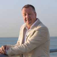 A man in a light-colored suit posing outdoors by a metal railing with the ocean in the background.