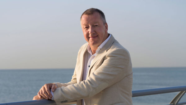 A man in a light-colored suit posing outdoors by a metal railing with the ocean in the background.
