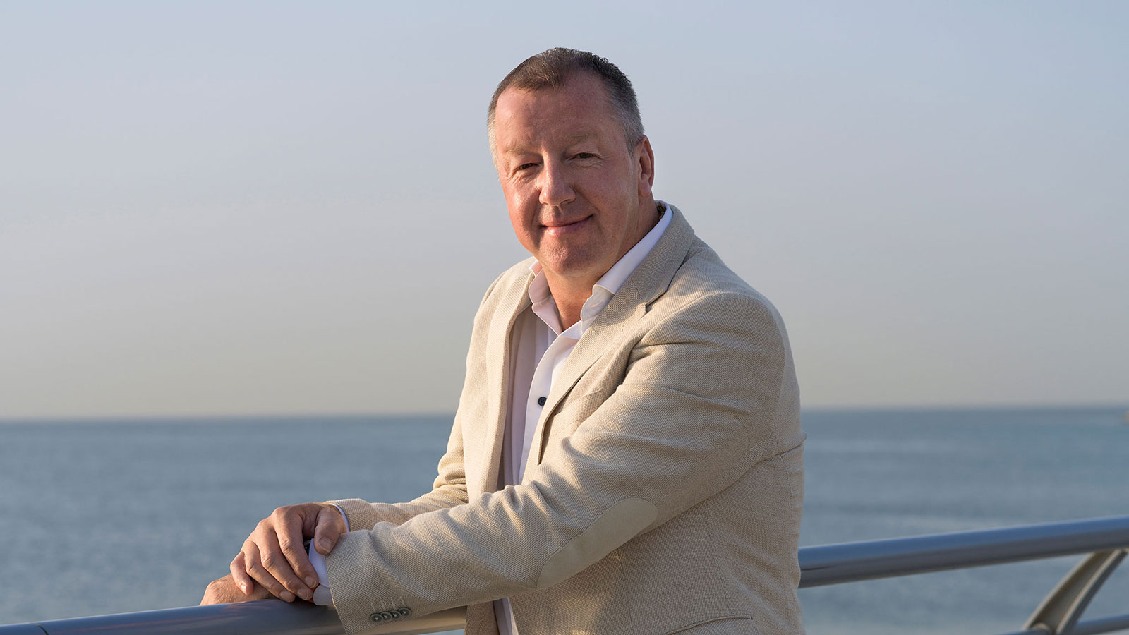 A man in a light-colored suit posing outdoors by a metal railing with the ocean in the background.