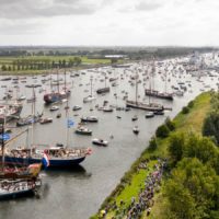 Aerial view of a river scene with numerous sailing ships and boats, some decorated with flags, navigating along the waterway. Crowds of people are gathered along the riverbank watching the maritime event.
