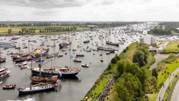 Aerial view of a river scene with numerous sailing ships and boats, some decorated with flags, navigating along the waterway. Crowds of people are gathered along the riverbank watching the maritime event.