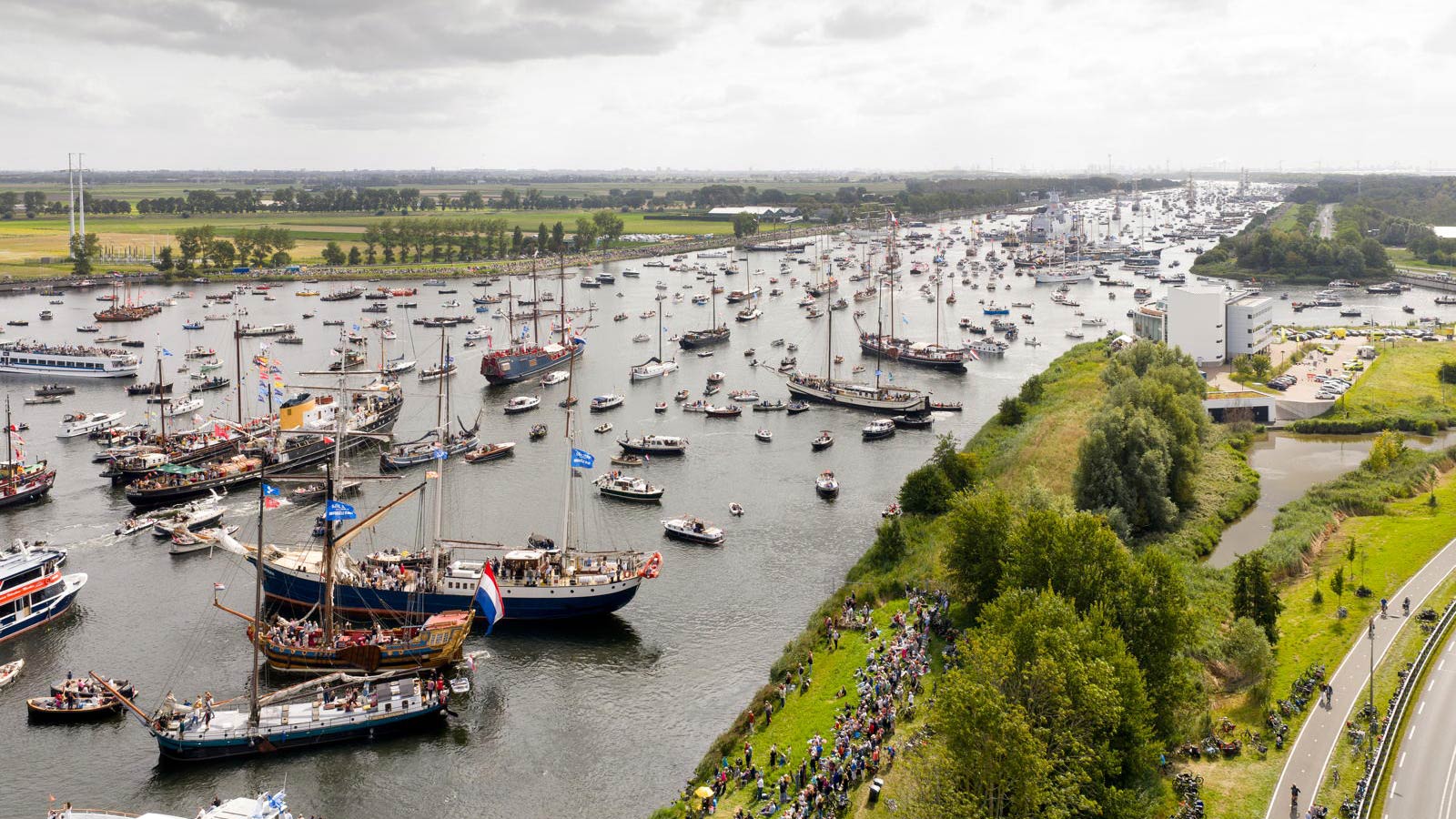 Aerial view of a river scene with numerous sailing ships and boats, some decorated with flags, navigating along the waterway. Crowds of people are gathered along the riverbank watching the maritime event.