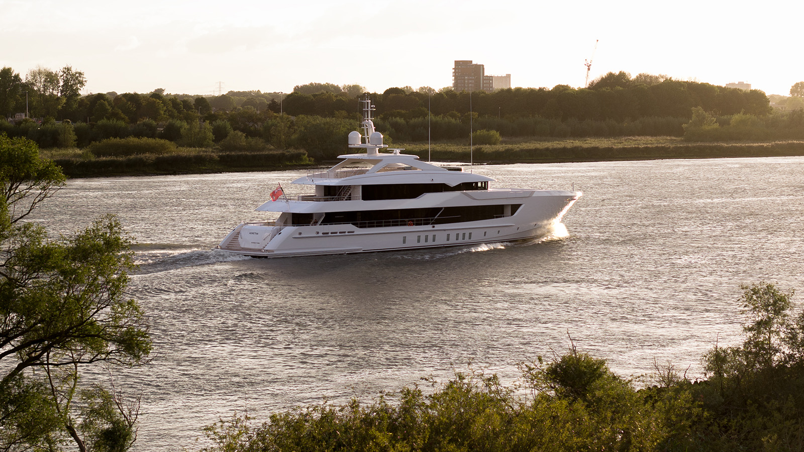 Luxury yacht cruising on a river with trees and buildings in the background, captured in warm evening light.