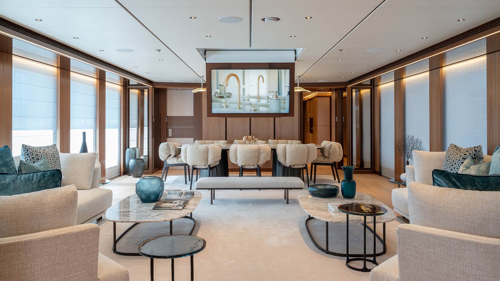 A luxurious living room with modern decor, featuring beige sofas, marble coffee tables, decorative pillows, and a large flat-screen TV on the wall, with large windows allowing natural light to enter.