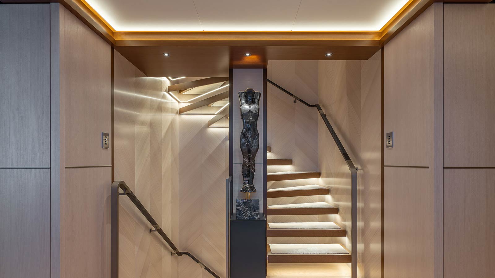 A modern, well-lit staircase with wooden panels and a decorative sculpture in the center dividing the stairs into two sections.