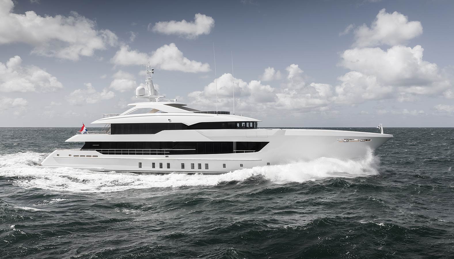 A large, luxurious white yacht cruising on the ocean, with waves breaking against its hull under a partly cloudy sky.