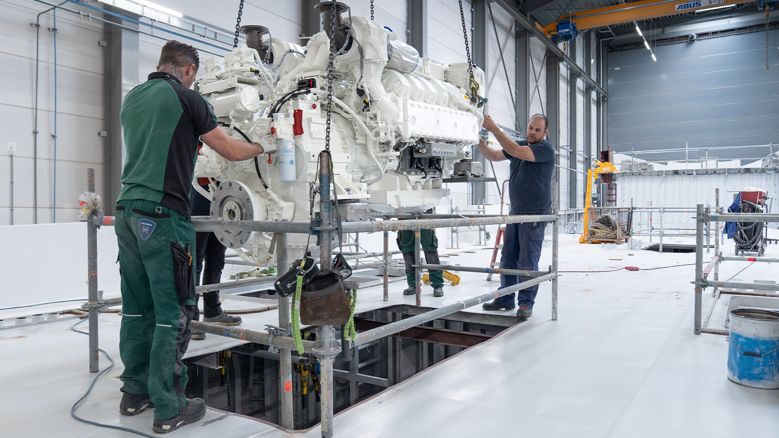 Two men working together to install a large, white engine suspended by a crane inside a spacious industrial facility, surrounded by scaffolding and construction equipment.