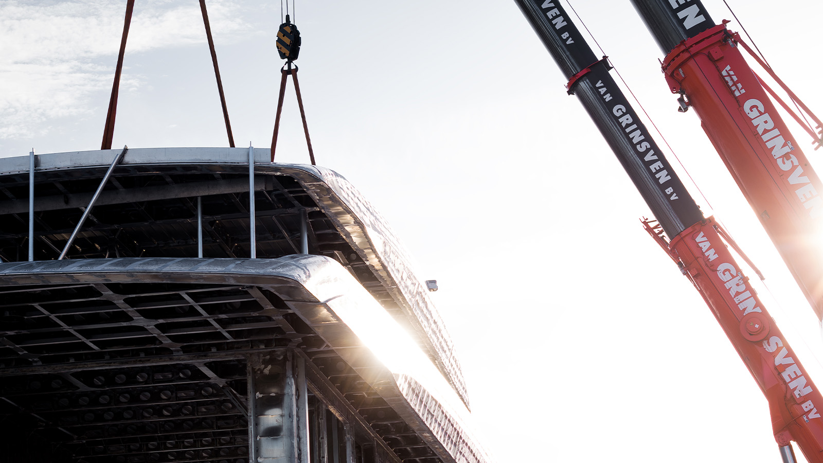 A crane lifting a large metallic structure with bright sunlight in the background, and two visible crane arms labeled "VAN GRINSVEN BV."