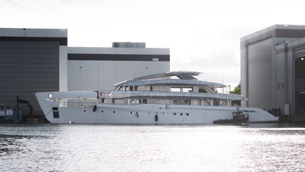 A large yacht under construction is docked between two industrial buildings at a shipyard. The vessel's hull appears complete, but the superstructure is still unfinished, showing exposed framework and metallic elements. The scene is set against a bright, cloudy sky.