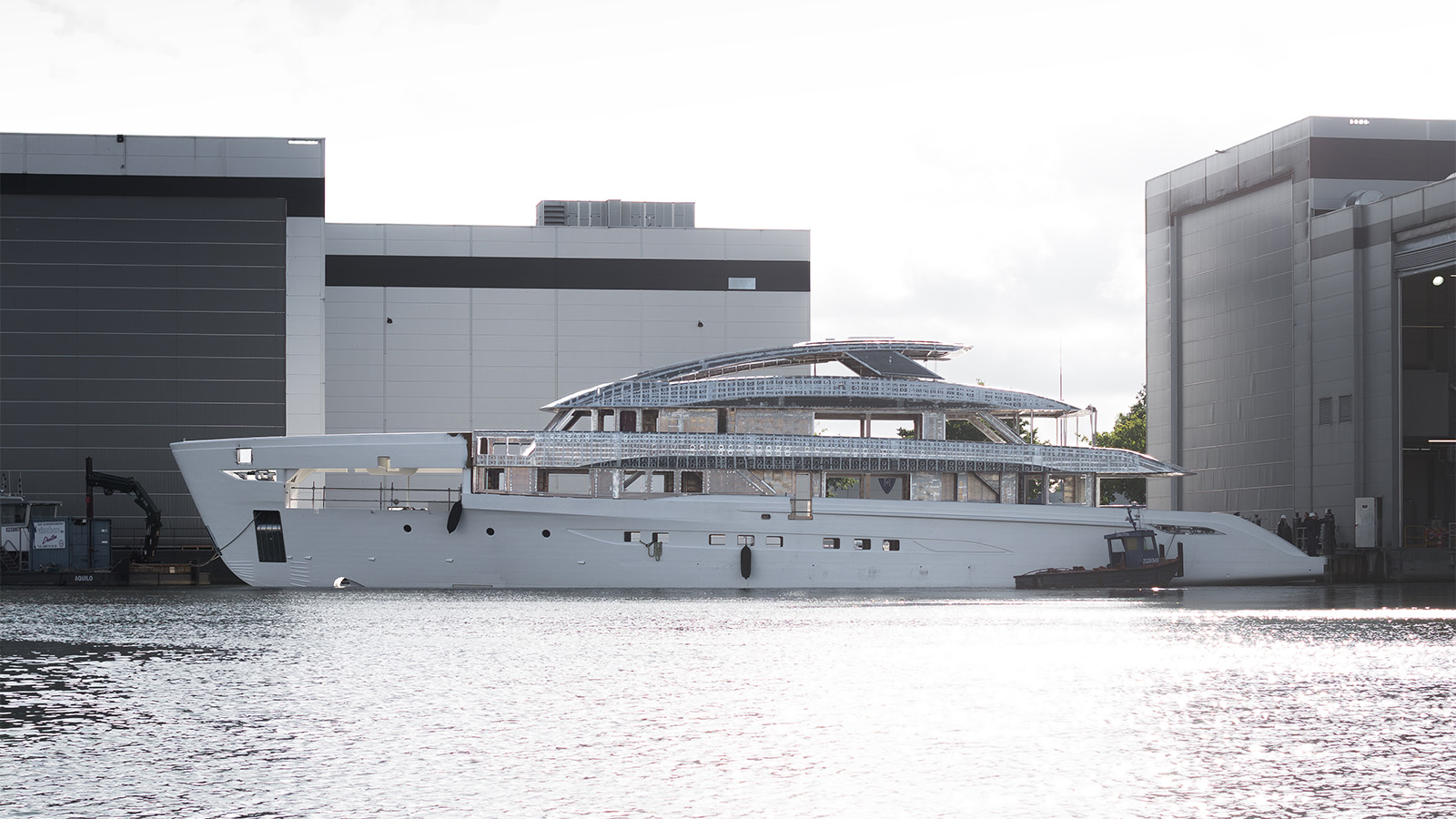 A large yacht under construction is docked between two industrial buildings at a shipyard. The vessel's hull appears complete, but the superstructure is still unfinished, showing exposed framework and metallic elements. The scene is set against a bright, cloudy sky.