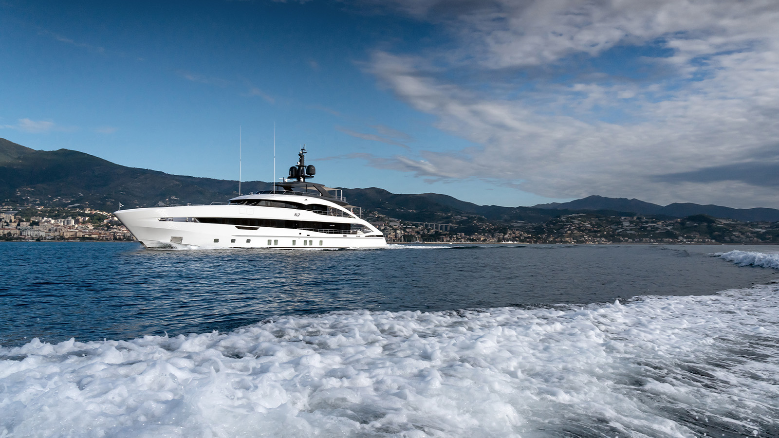 A luxurious white yacht sails across a calm sea with a hilly coastline in the background and a trail of wake in the foreground under a partly cloudy sky.